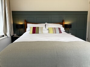 A neatly made bed with white linens and multiple pillows, a wooden headboard, bedside lamps, and a window with curtains.