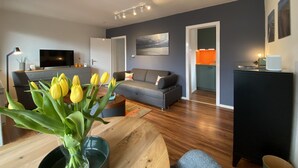 Ein modernes Wohnzimmer mit einem grauen Sofa, einem Holztisch mit einer Vase voller gelber Tulpen, einem Flachbildfernseher und einem Bild an der Wand.