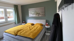 Ein Schlafzimmer mit Bett, Nachttisch, Kleiderhaken, einem Gemälde und einem Fenster.