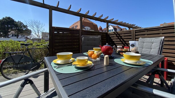 Ein Essbereich im Freien mit einem für zwei Personen gedecktem Tisch, umgeben von einer Holzpergola.