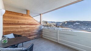 En balkong med en trävägg, ett bord och stolar, med utsikt över ett snöklätt berglandskap.