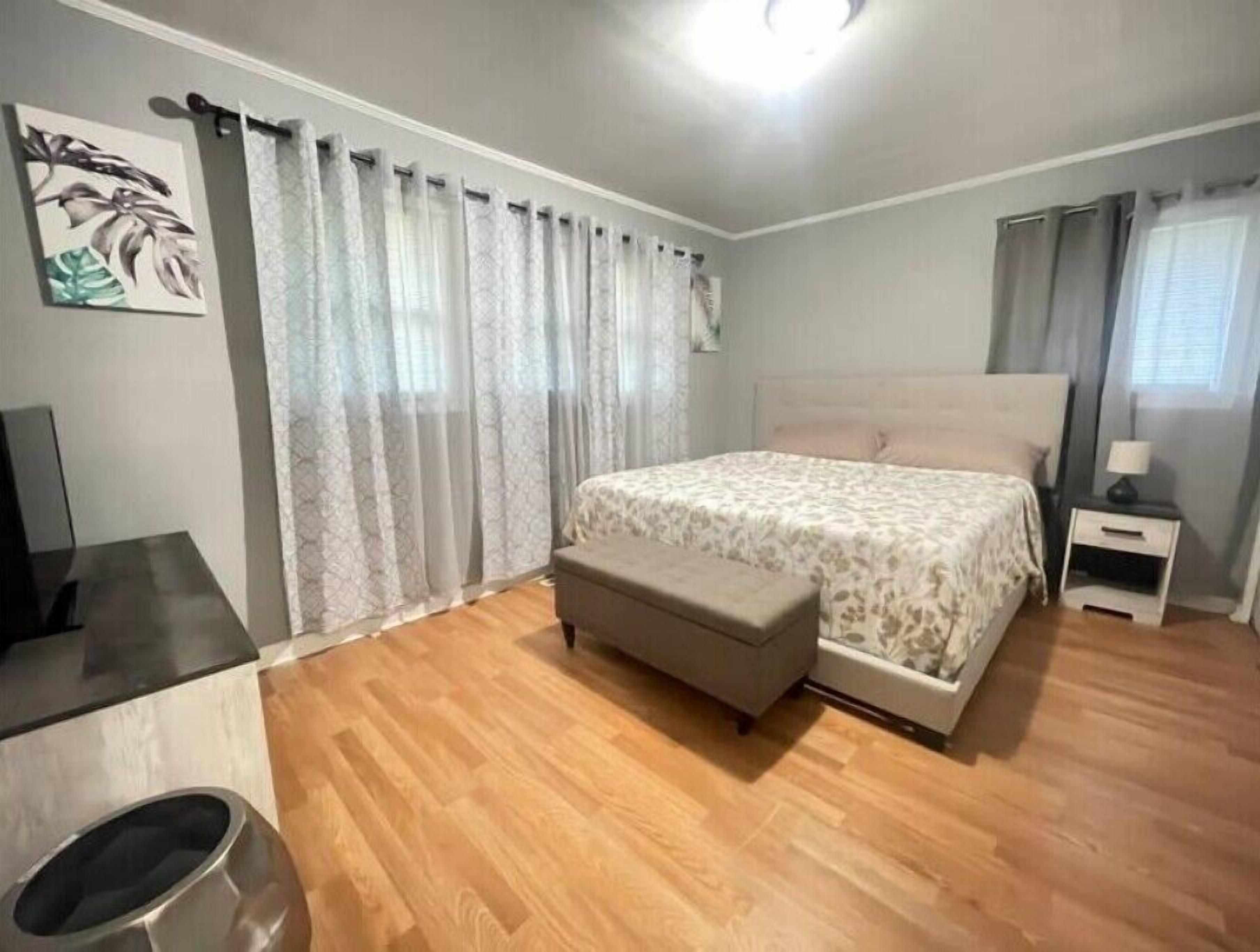 Une chambre à coucher comprenant un lit, une table de chevet, un banc, un téléviseur et une fenêtre avec des rideaux.