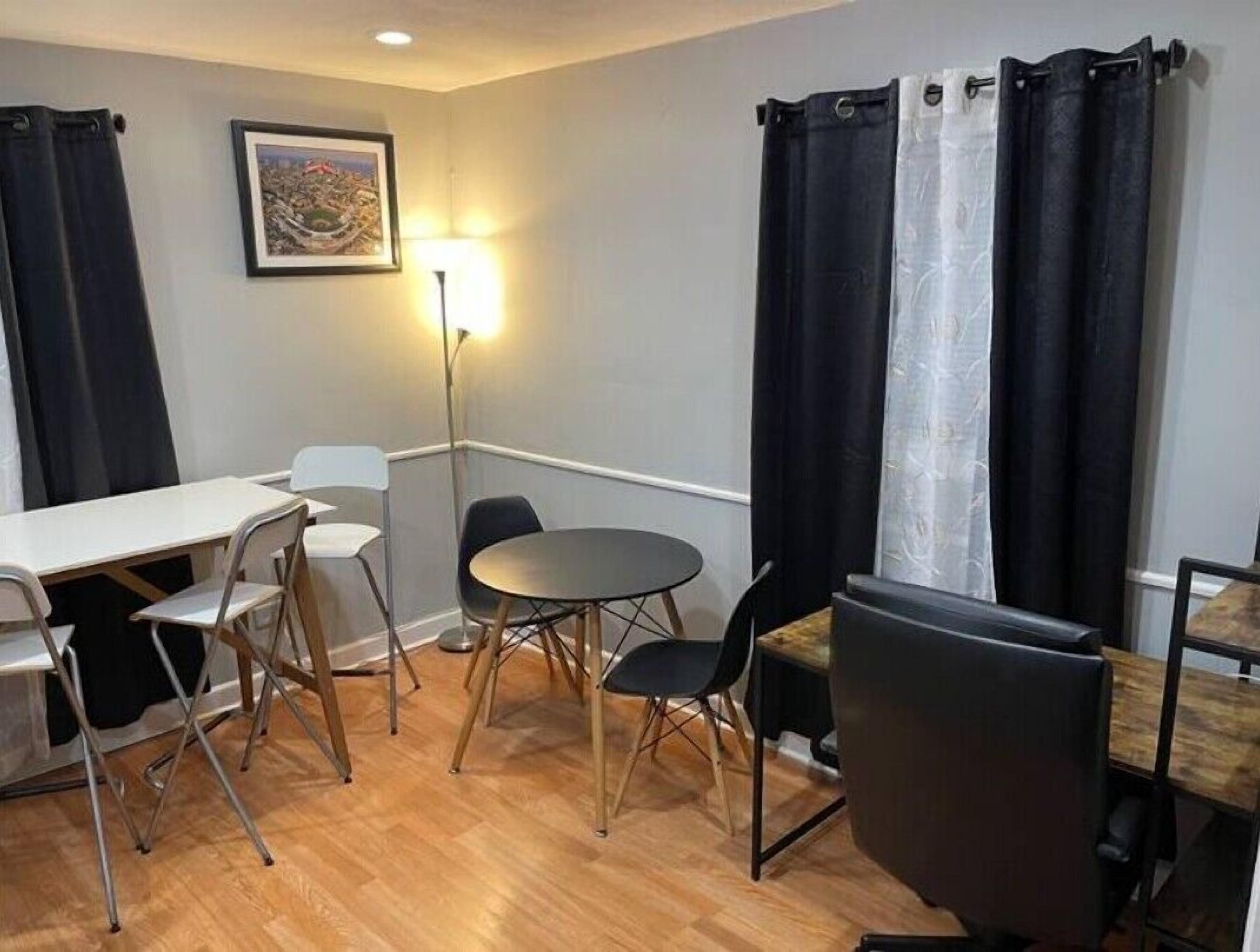 A small, furnished room with a wooden floor, a white table, two chairs, a round table, and a black chair.