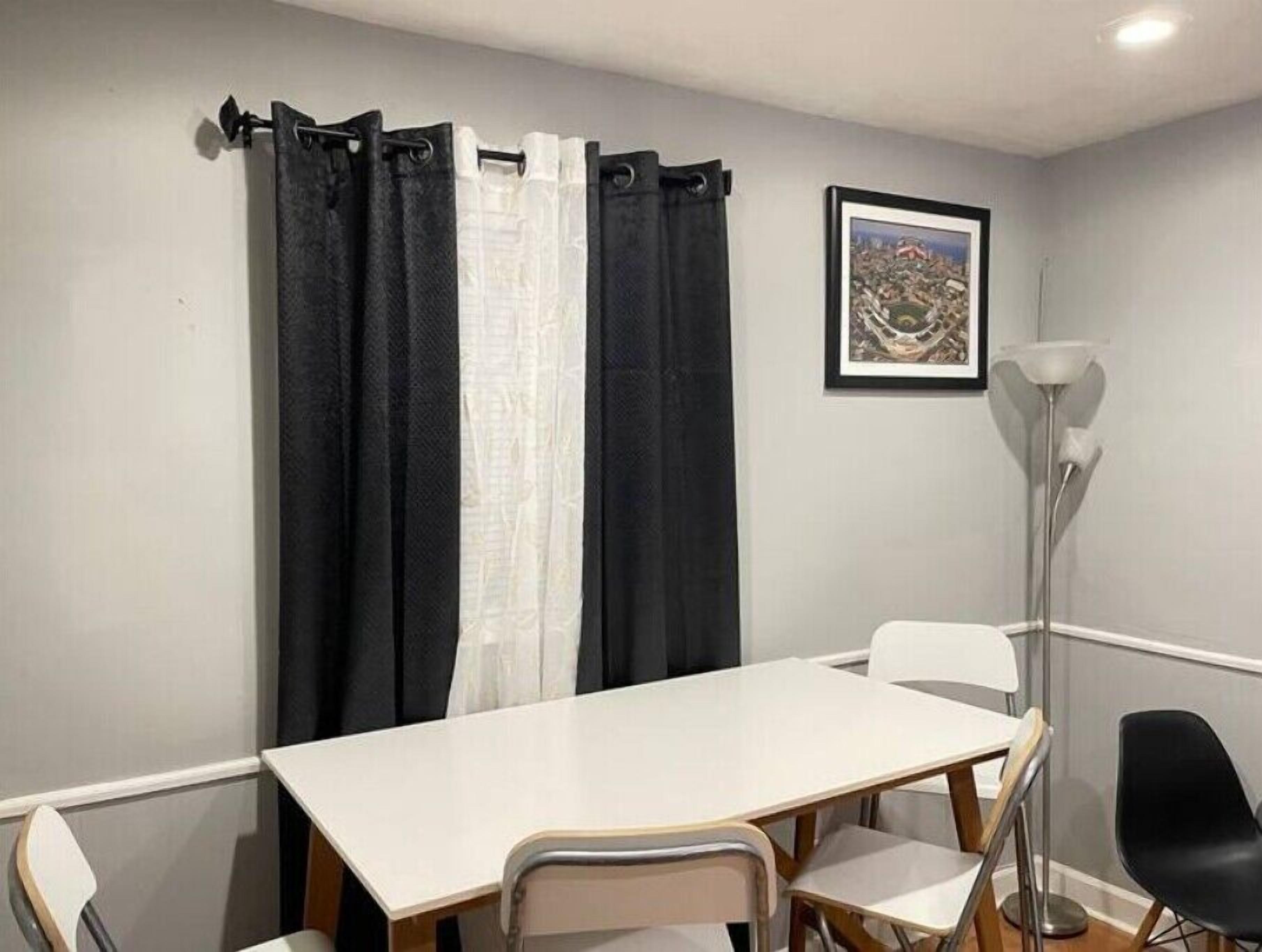 A room with a white table, four chairs, black and white curtains, a framed picture on the wall, and a floor lamp.