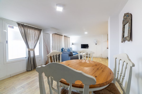 A modern living room with a round wooden dining table, white chairs, a blue sofa, and a TV mounted on the wall. - AlmeriaSuite Mojacar Playa (Mojacar)