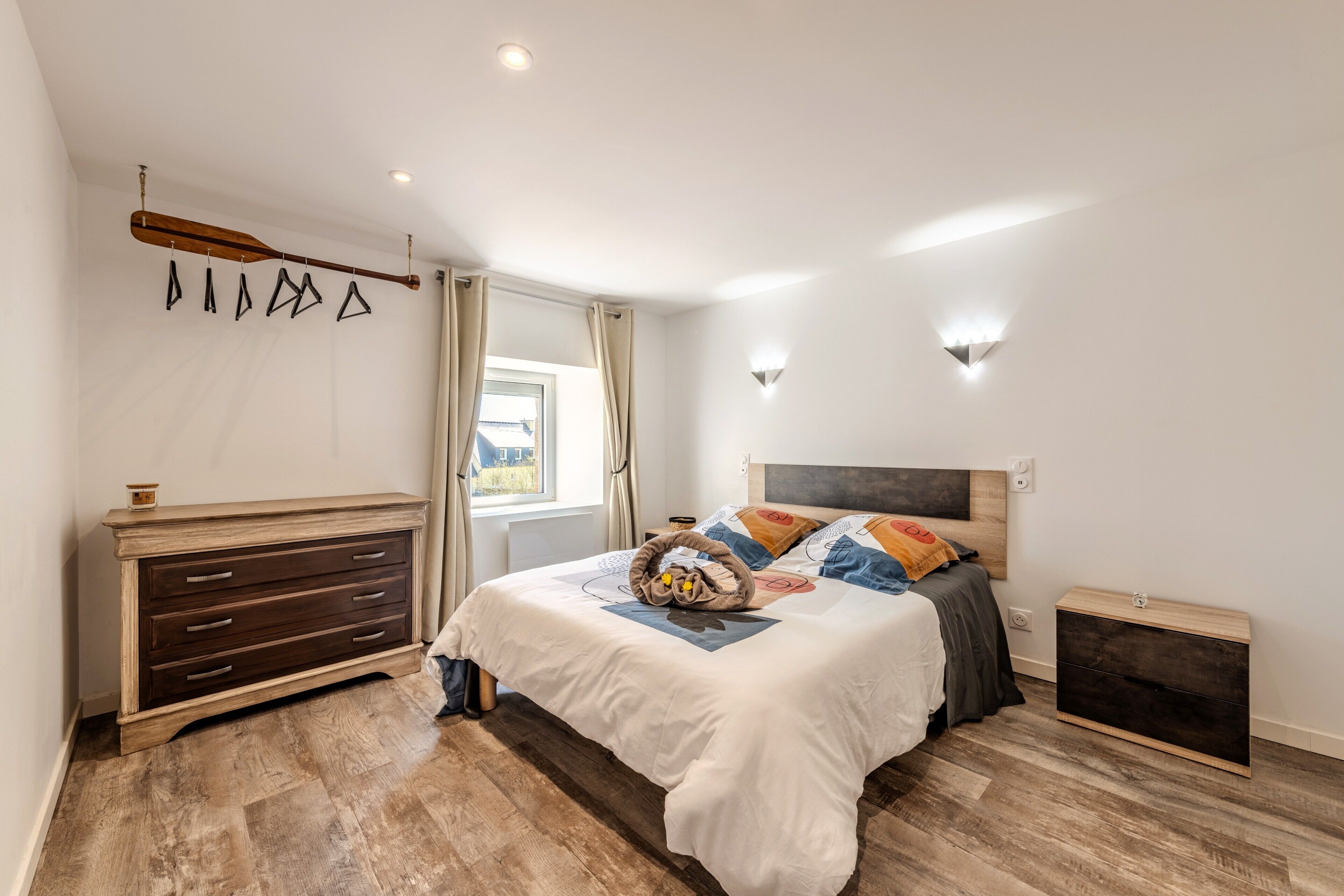 Een slaapkamer met een houten dressoir, een bed met meerdere kussens, een hoofdbord en een raam met gordijnen.