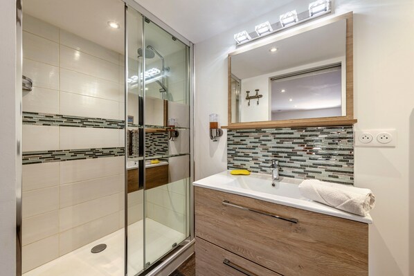 A modern bathroom with a glass shower enclosure, a white sink with a wooden vanity, and a mirror with a tiled backsplash.