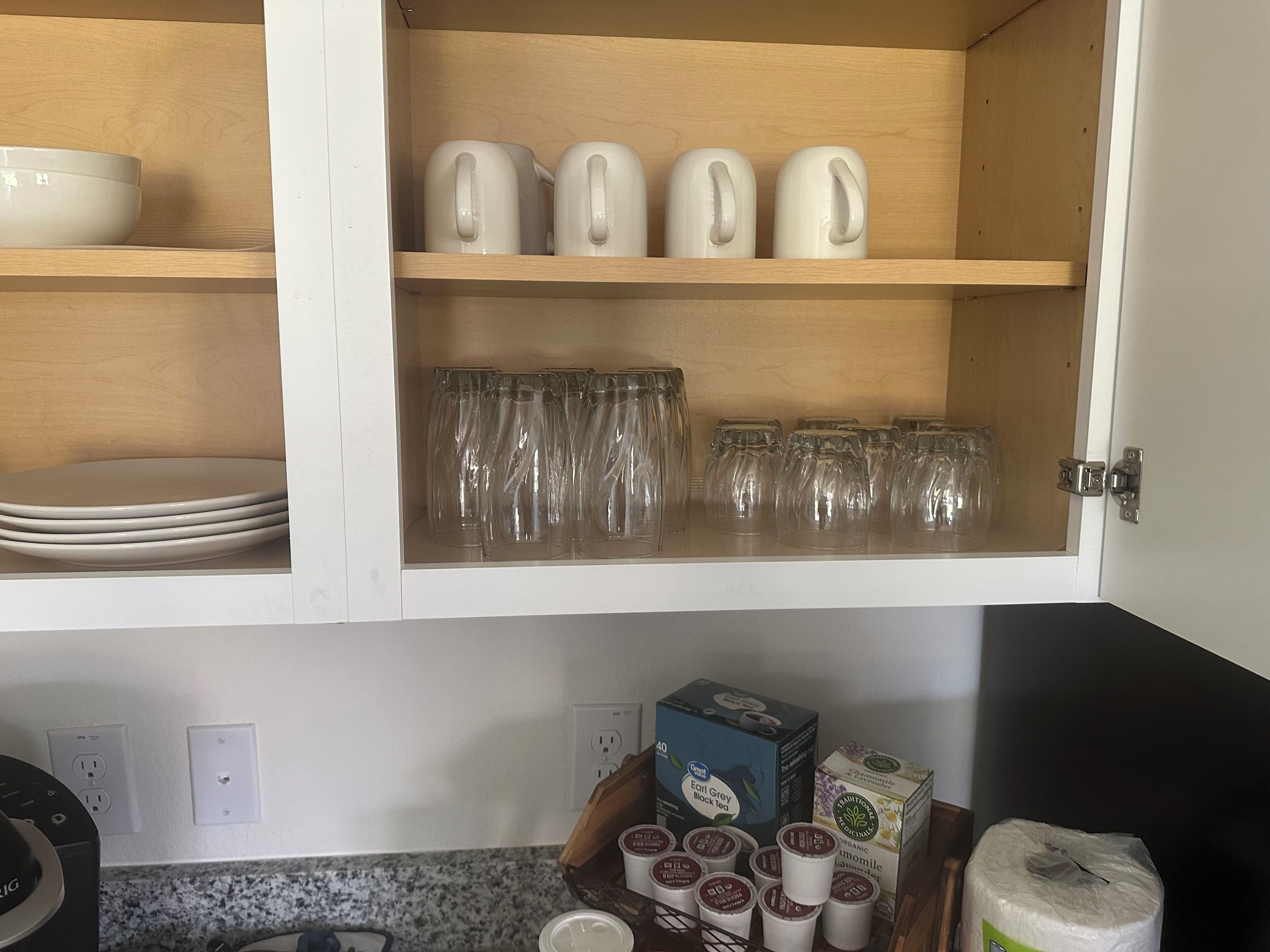 A kitchen cabinet with white plates, clear glasses, and white teapots.