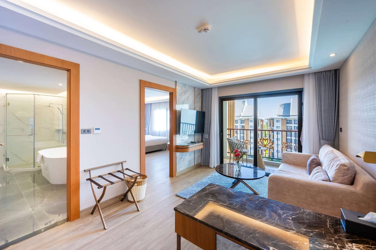 Moderni hotellihuone, jossa on lasinen suihkukaappi, sohva, televisio ja parveke, jolta on näkymä kaupunkiin.