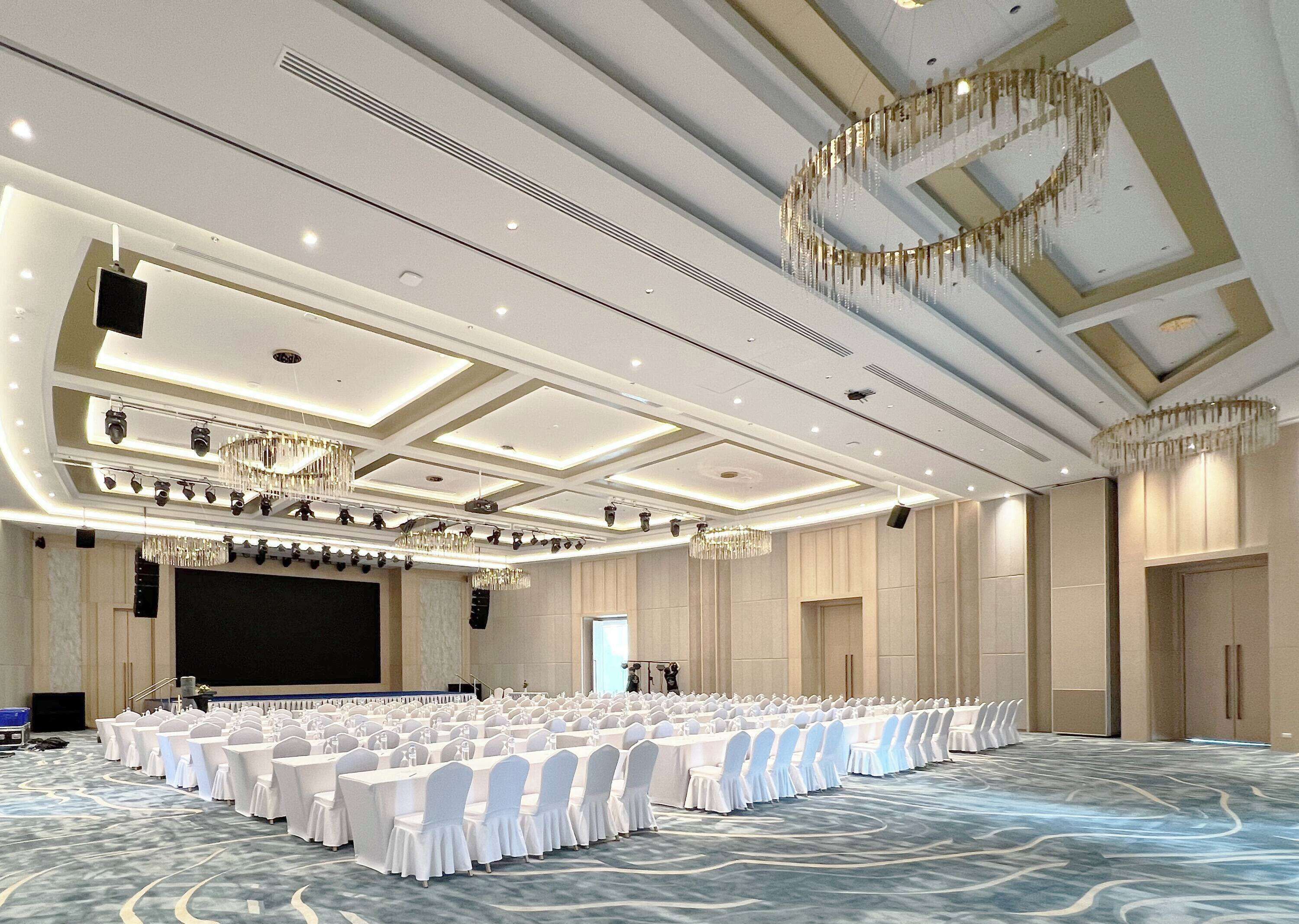 A large conference room with rows of tables and chairs, a stage, and a high ceiling with chandeliers.
