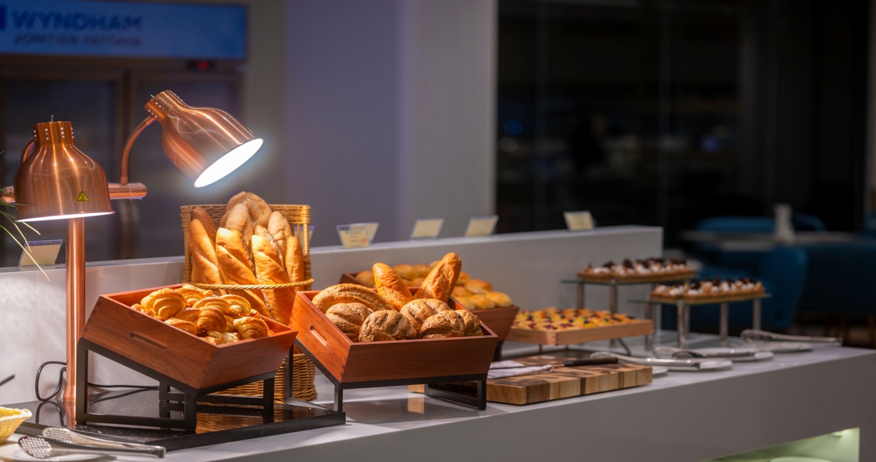Um buffet com diversos produtos de panificação e pastelaria, um candeeiro de cobre e uma placa ao fundo.