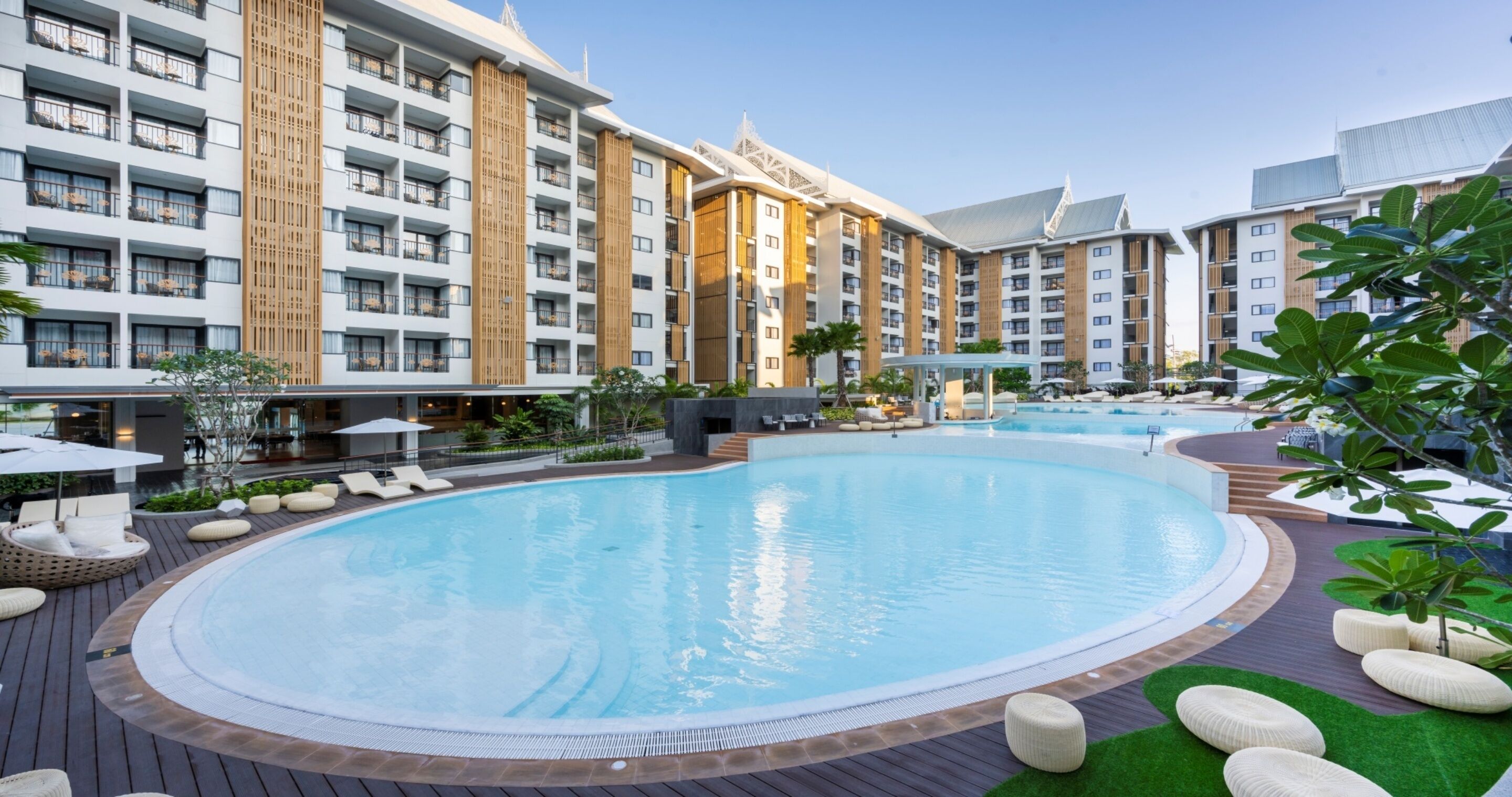 A hotel with a large swimming pool surrounded by lounge areas and greenery.