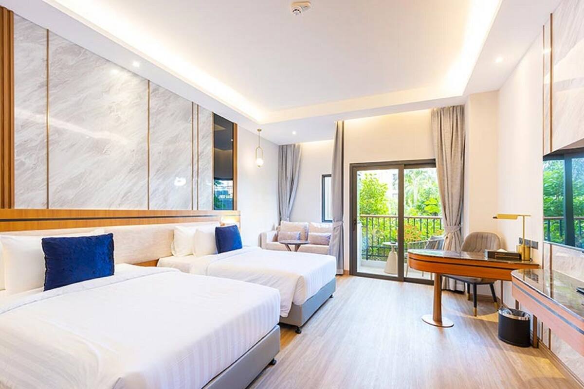 A hotel room with two beds, a desk, and a balcony with a view of greenery.