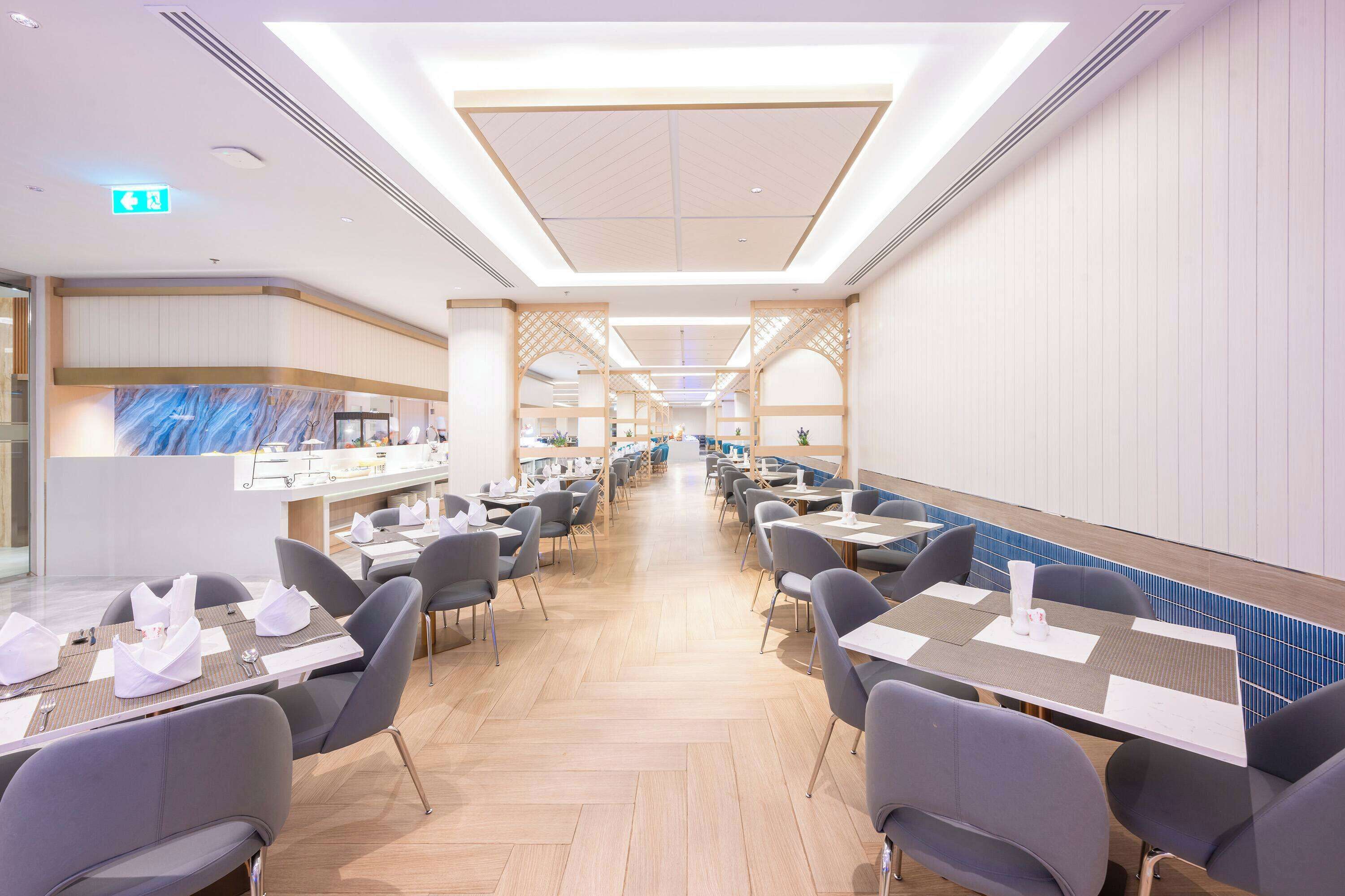 Amplia zona de comedor de hotel con múltiples mesas y sillas, un mostrador de recepción y un diseño de techo moderno.