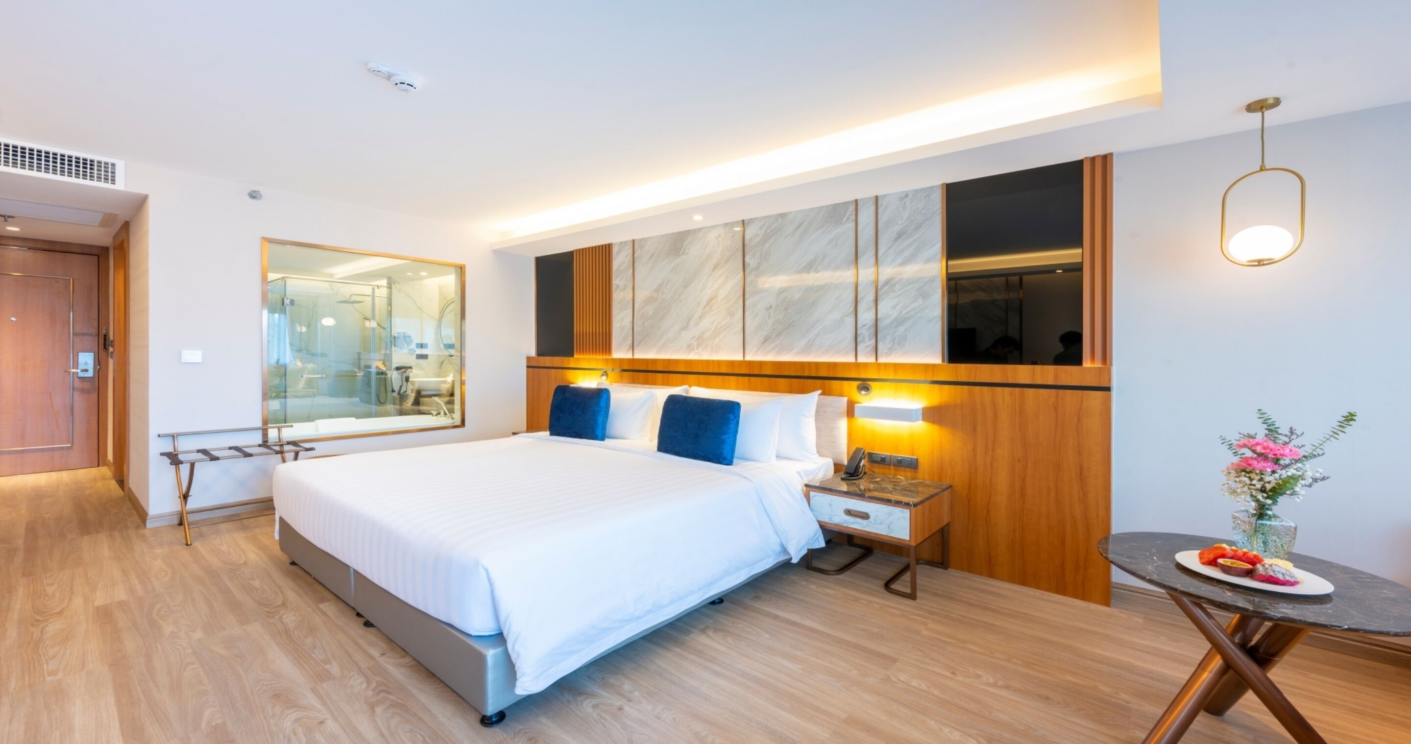 A modern hotel room with a large bed, bedside tables, a mirror, a small table with a vase, and a wooden floor.