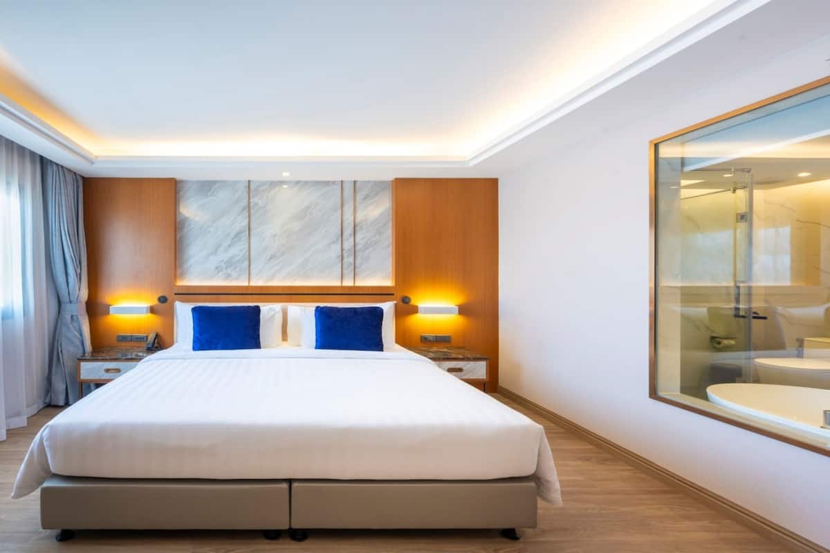 Moderni hotellihuone, jossa on suuri sänky, puinen pääty, kaksi yöpöytää valaisimineen, suuri peili ja näkymä kylpyhuoneeseen.