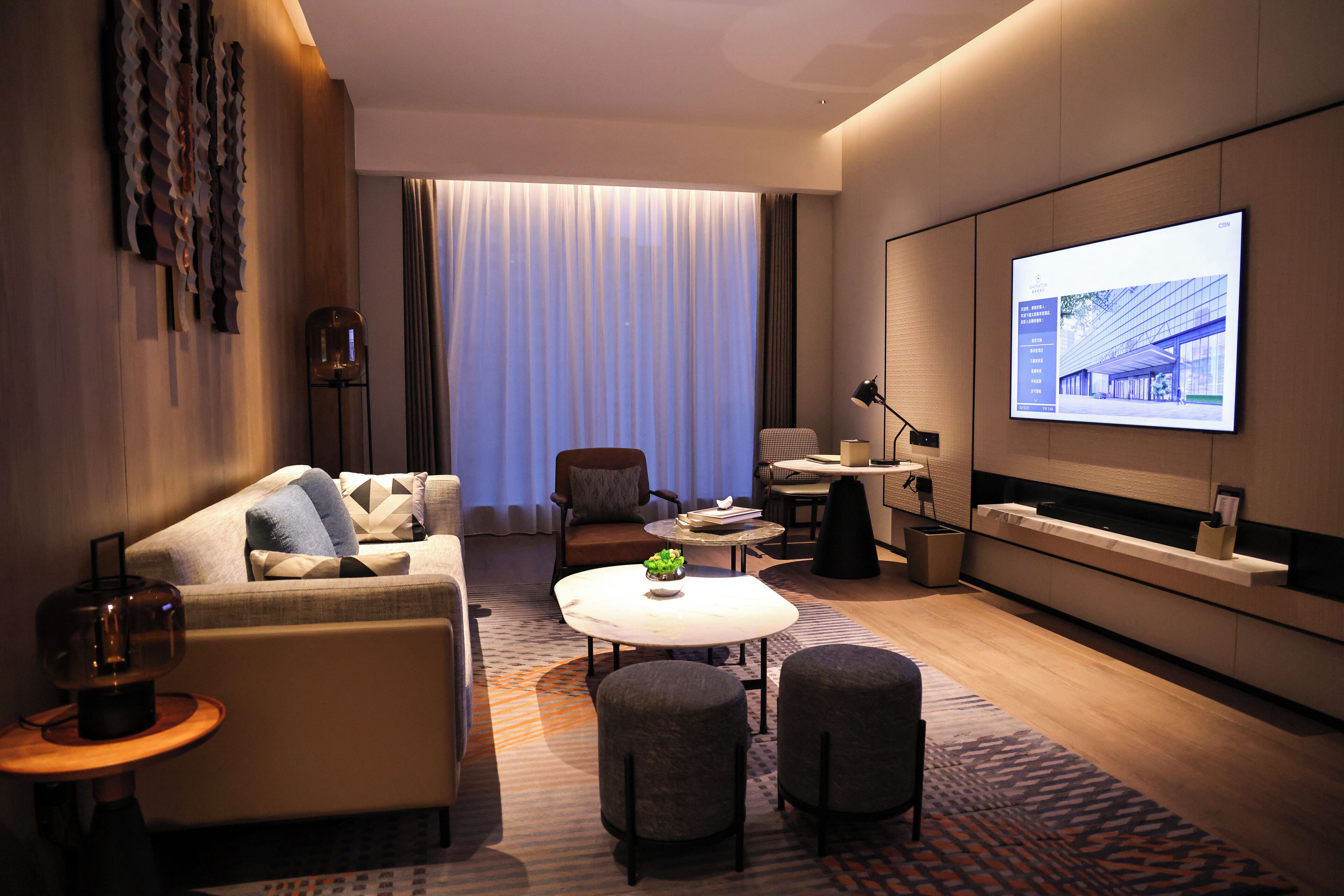 A modern hotel room with a sofa, armchair, ottoman, coffee table, and a large TV screen displaying a building image.