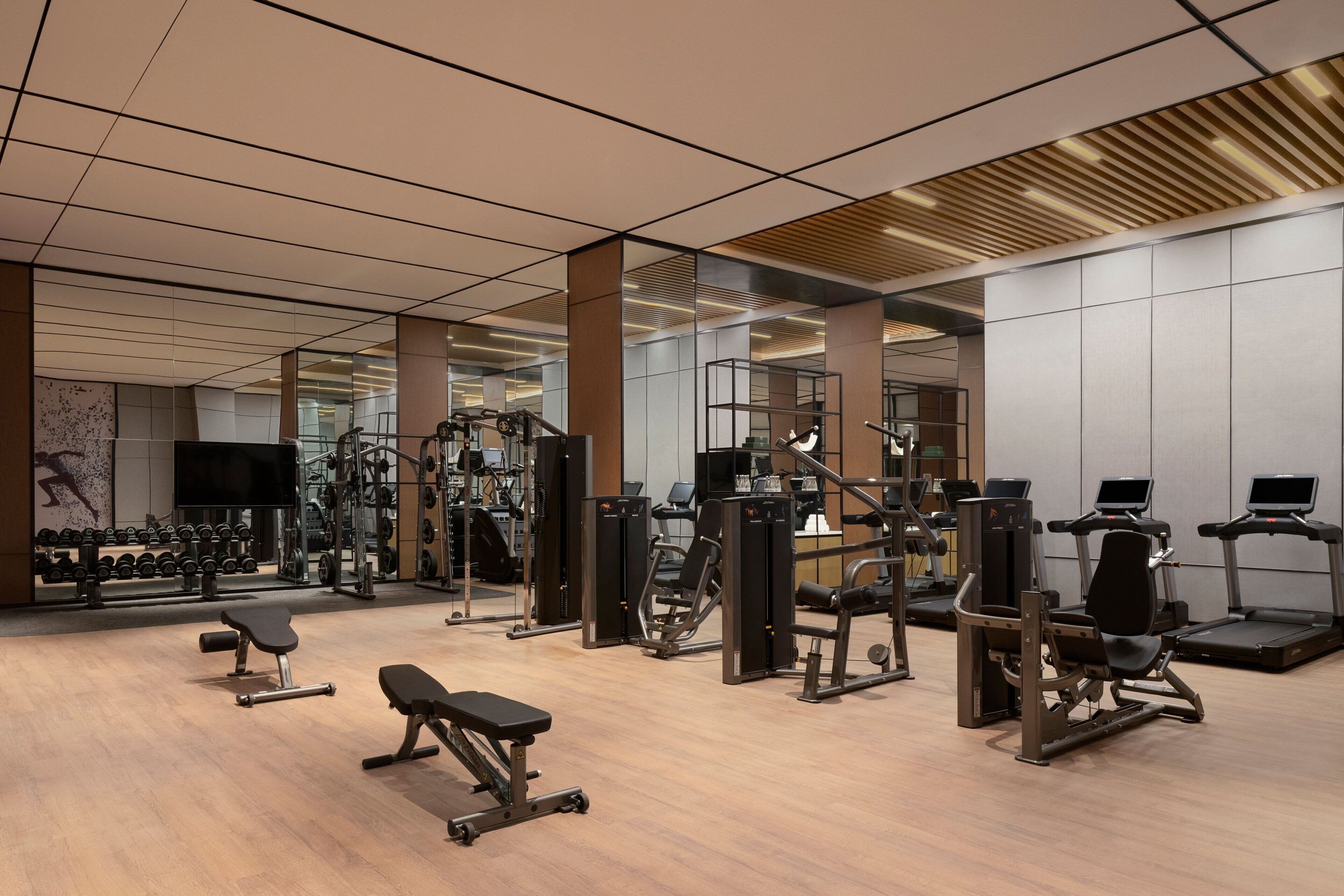 A modern gym with various exercise equipment, large mirrors, and a wooden floor.
