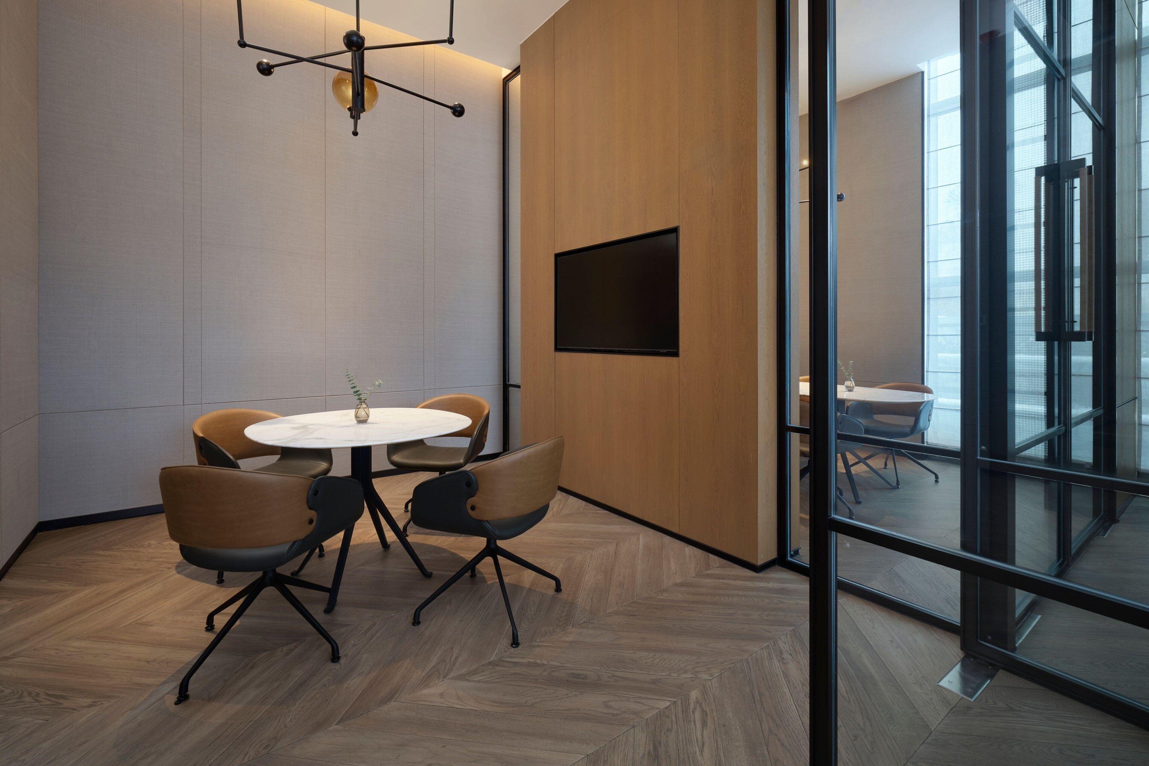 A modern meeting room with a round table, four chairs, a mounted TV, and a large glass wall.