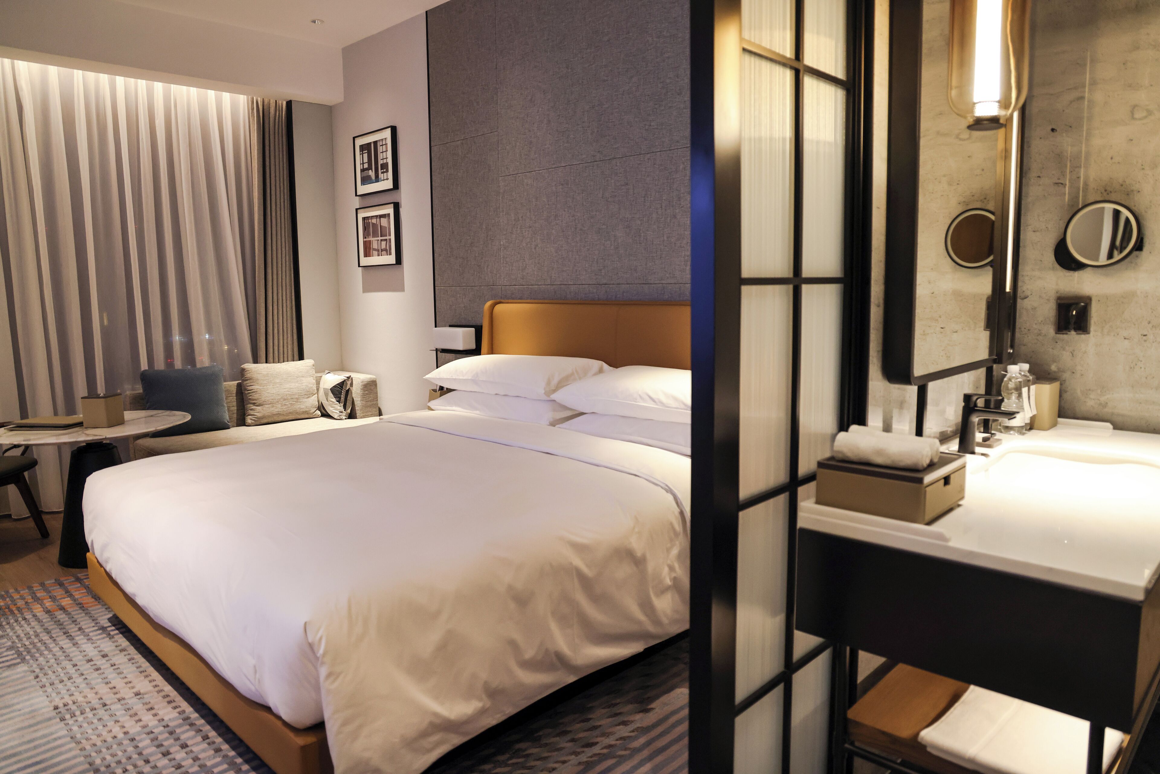 A hotel room with a large bed, a bedside table, a small round table, a sofa, and a bathroom with a sink and mirror.