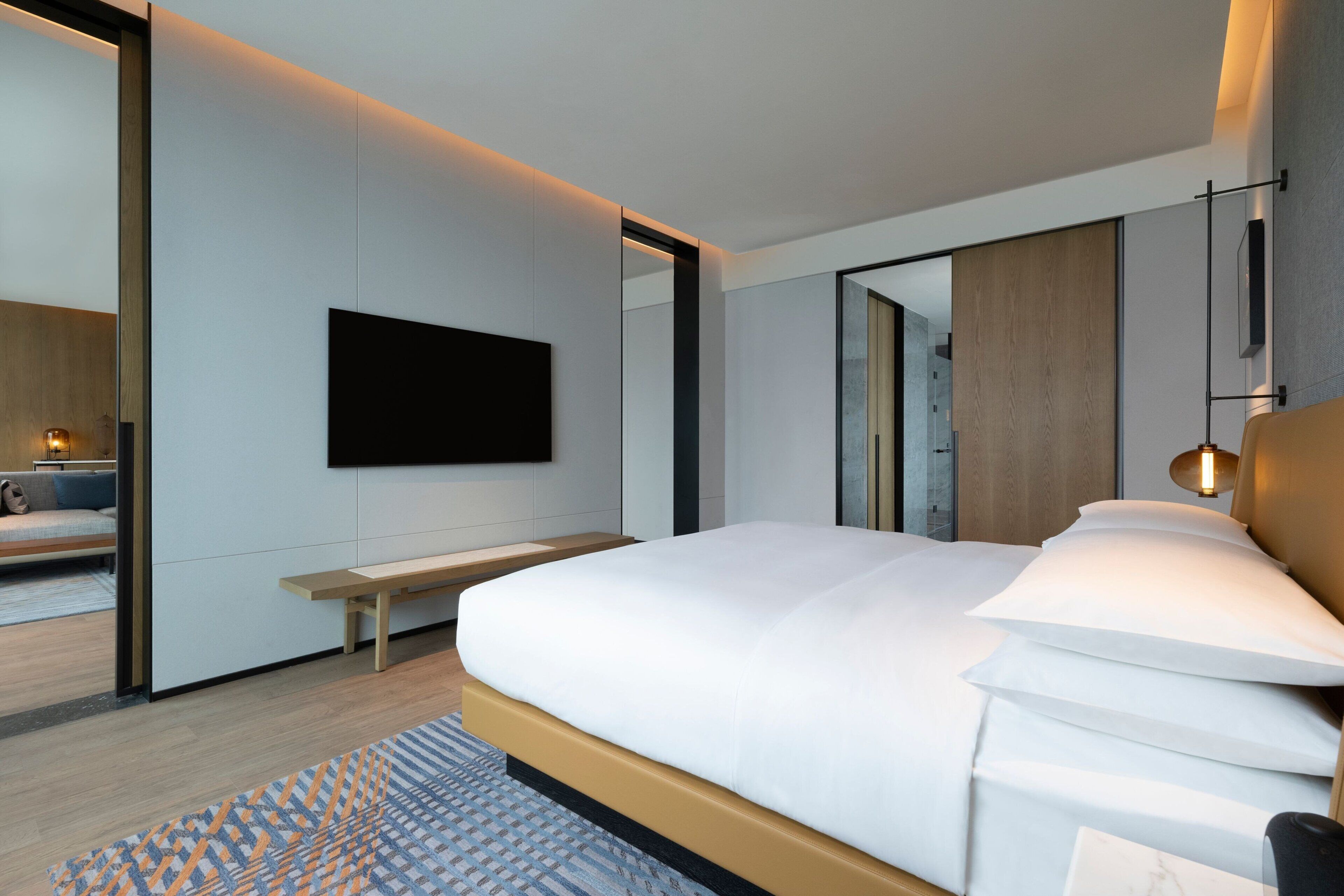 A modern bedroom with a large bed, a flat-screen TV mounted on the wall, a bedside table, and a large mirror.