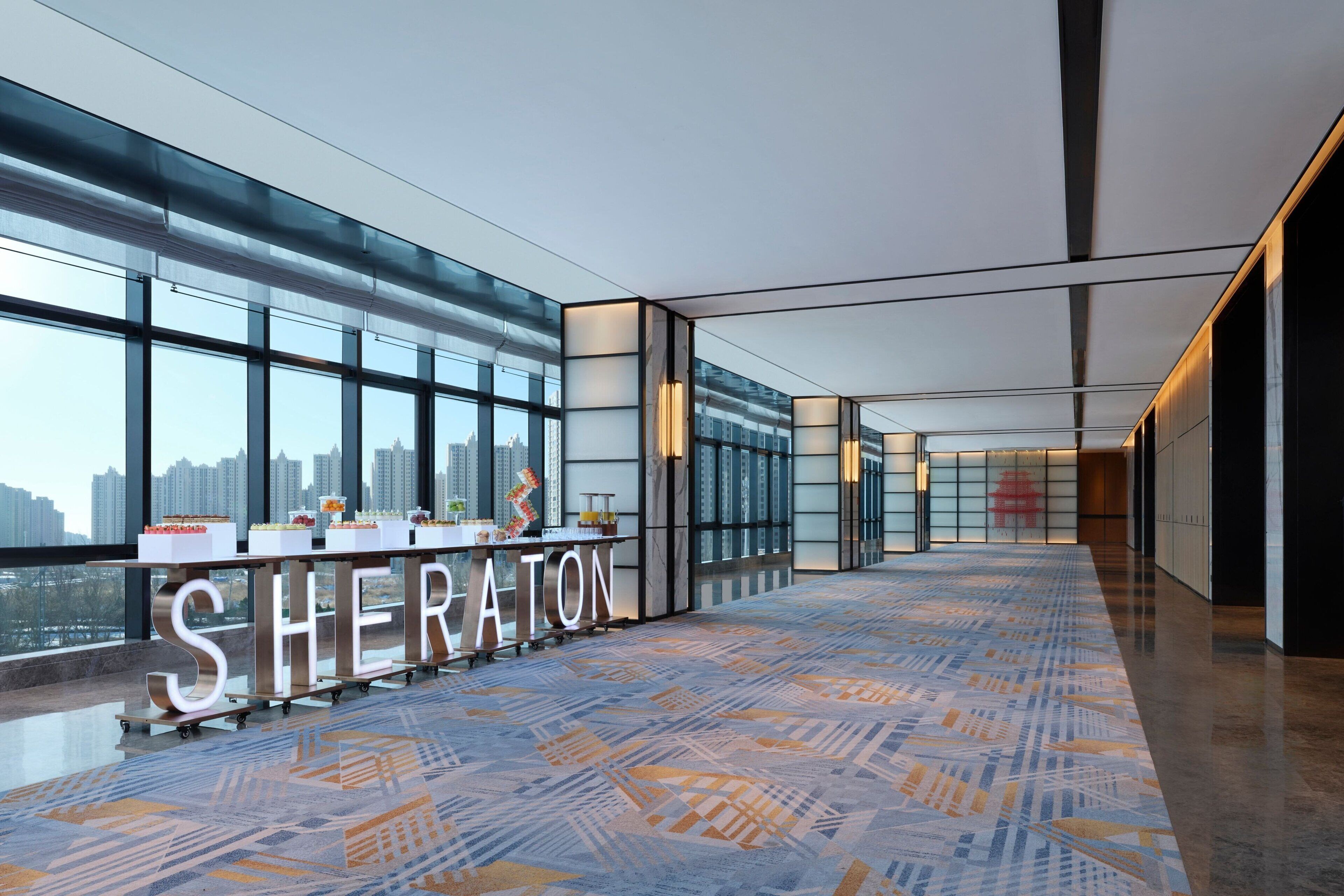 Un hall d’hôtel spacieux au design moderne, avec un long comptoir d’accueil portant l’inscription « SHERATON » et de grandes fenêtres offrant une vue sur la ville.