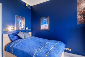 A bedroom with a blue bedspread, two framed pictures on the wall, and a nightstand with a lamp.