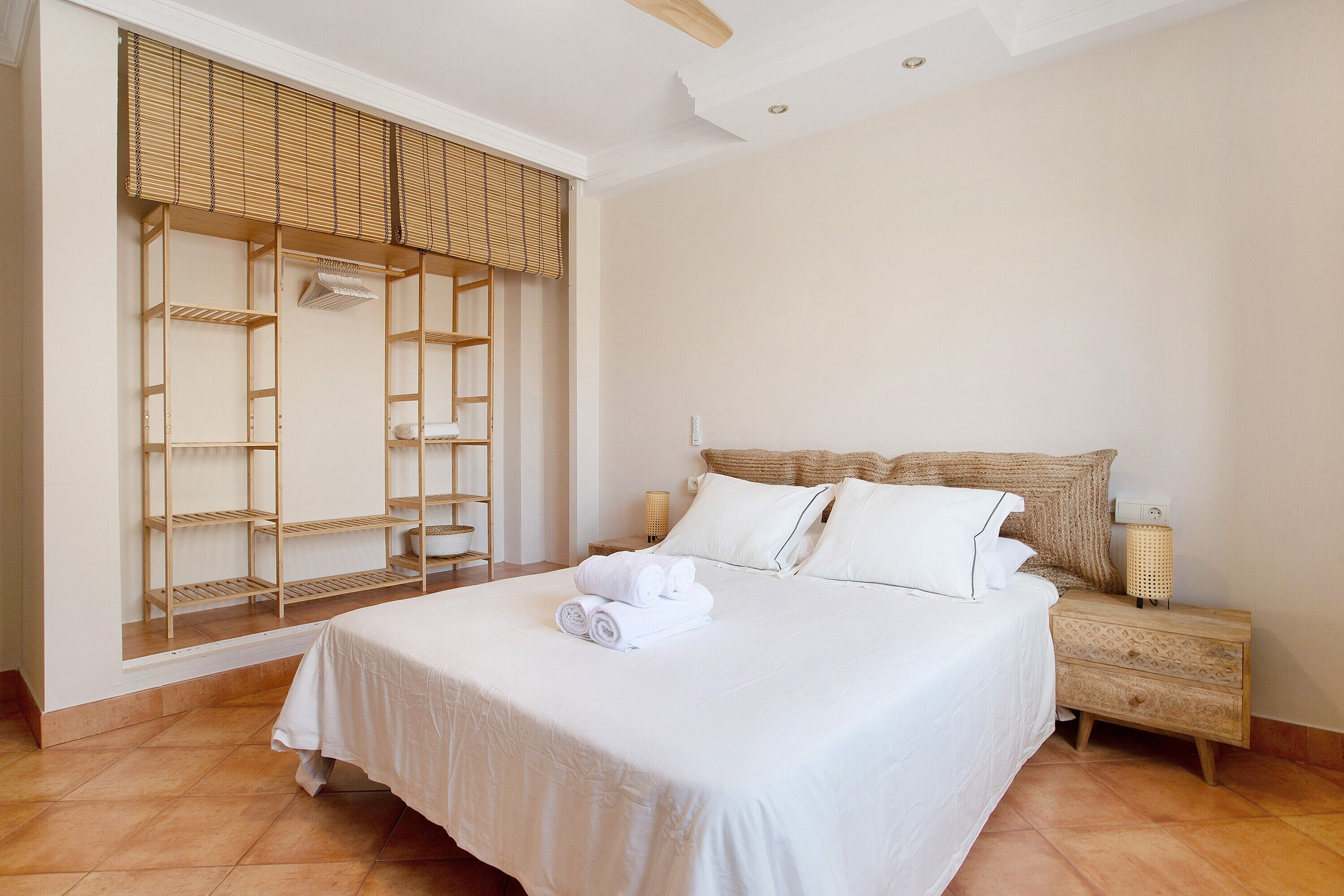 Un letto rifatto con precisione, una coperta bianca, un comodino in legno con lampada e un armadio a muro con ripiani.