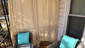 Varanda coberta com duas cadeiras de vime, cada uma com almofada, uma mesa de madeira pequena e um tapete estampado.