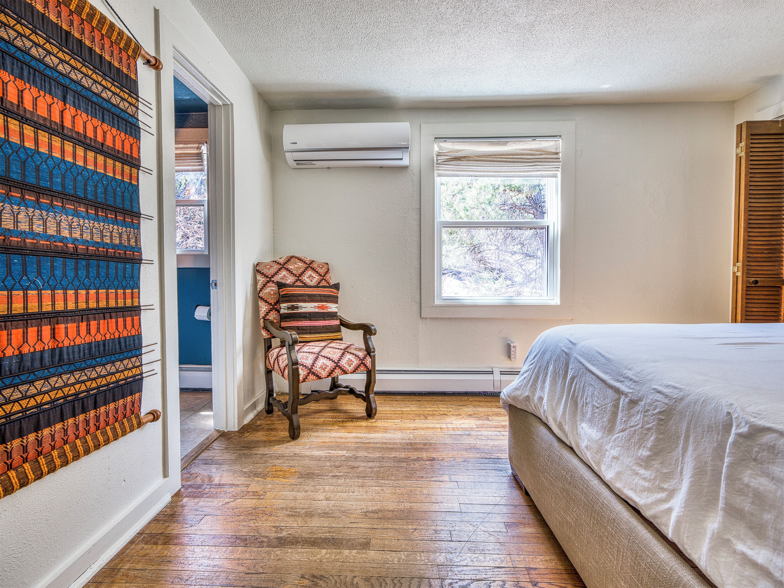 Une chambre avec un plancher en bois, un lit recouvert de draps blancs, une chaise agrémentée de coussins à motifs, une fenêtre avec des stores et un climatiseur fixé au mur.