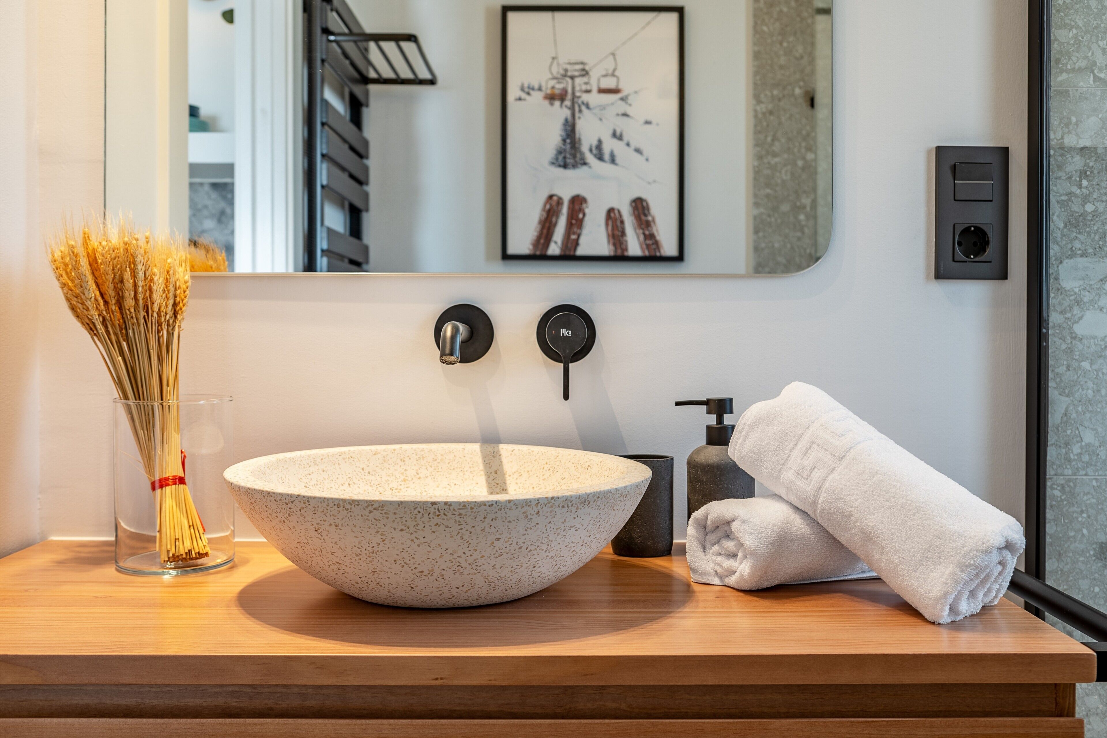 Une salle de bain avec un grand lavabo en forme de coupe, un vase en verre contenant des tiges de roseau, une serviette, un tableau encadré et un miroir.
