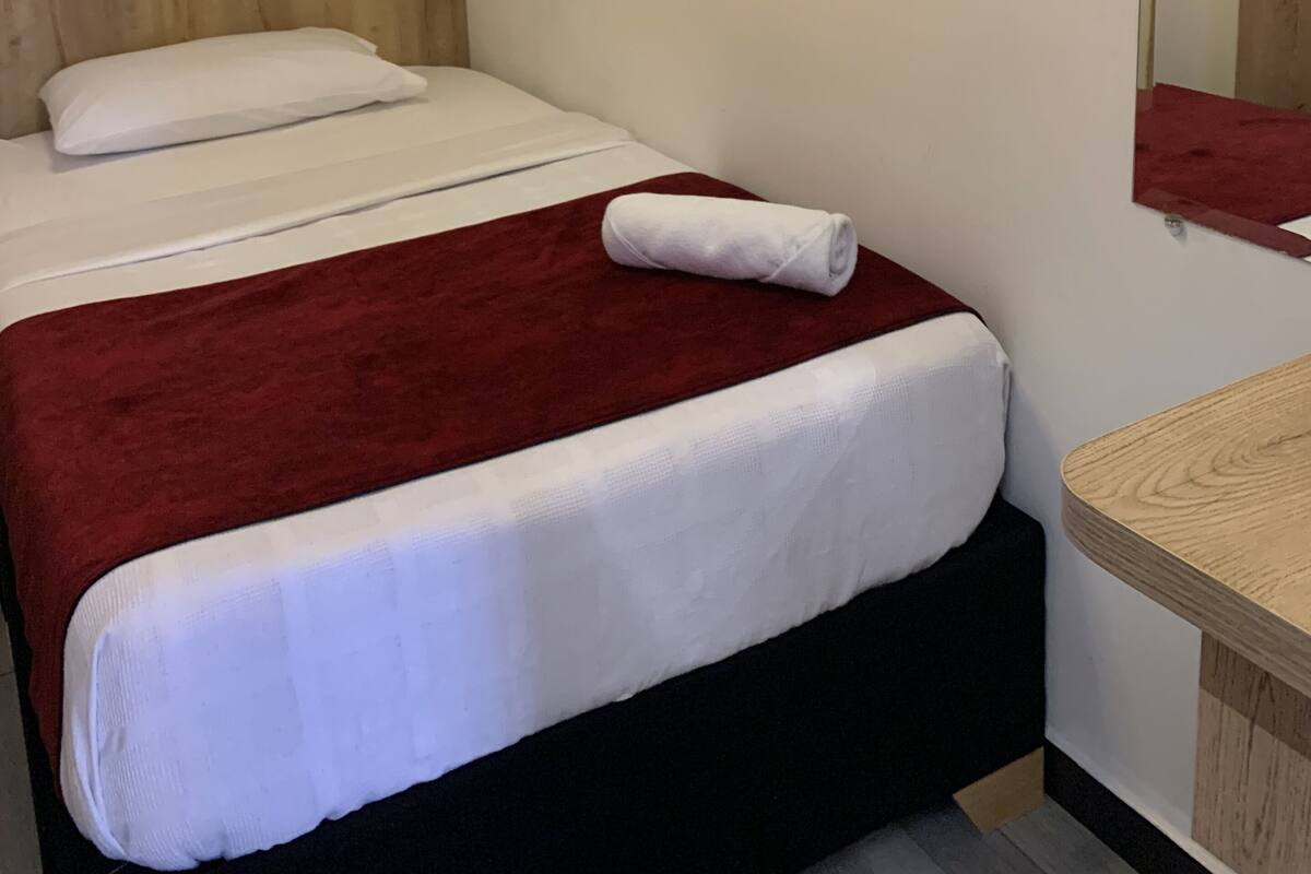 Una cama individual con cabecera de madera, ropa de cama blanca y una manta roja. Una mesita de noche de madera pequeña con un espejo y una toalla roja encima.