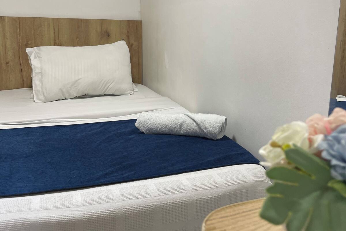 Una cama bien hecha con una manta azul y ropa de cama blanca, un cabecero de madera y una mesita con una planta.