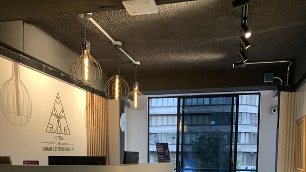 Zona de recepción de hotel con diseño moderno, que incluye un mostrador, iluminación colgante y vistas al exterior a través de grandes puertas de cristal.