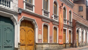 Vista de la calle con un edificio que tiene puertas de madera grandes, fachada de piedra y un cartel rojo.