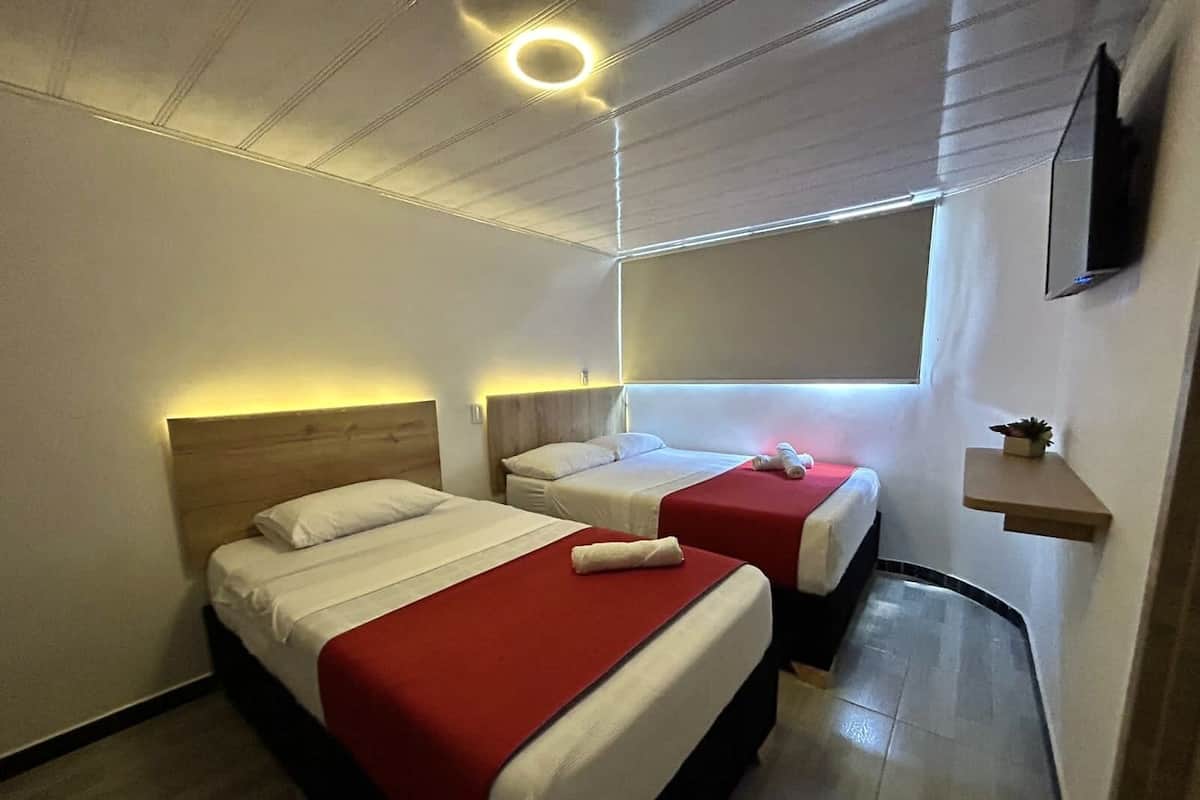Habitación de hotel con dos camas, cada una con cabecera de madera, ropa de cama blanca y colchas rojas. Se observa una pequeña estantería con una planta y un televisor de pantalla plana montado en la pared.