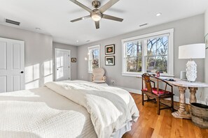 Ein Schlafzimmer mit einem Bett, einem Schreibtisch, einem Stuhl, einem Deckenventilator und großen Fenstern.