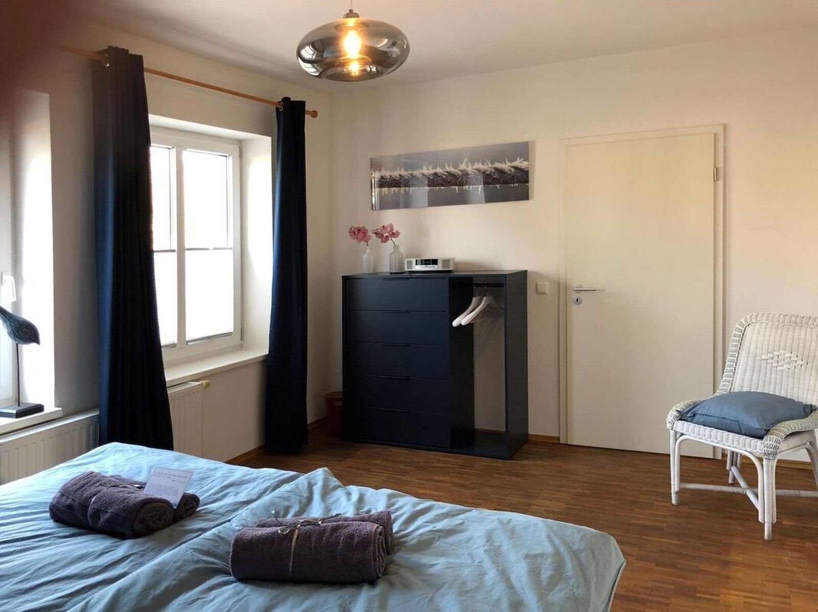 Une chambre d’hôtel comprenant un lit, une commode, une chaise, une fenêtre avec des rideaux et un luminaire suspendu.
