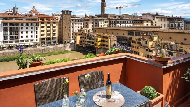 Una terraza con una mesa, sillas, una botella y vasos, con vistas a un centro histórico con edificios y un río.