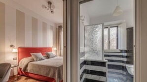 Hotelkamer met een rode hoofdbord, een houten visgraatvloer, een groot bed, een zithoek met een fauteuil, en uitzicht op een stenen muur en balkon.
