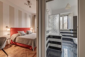 A hotel room with a red headboard, a wooden herringbone floor, a large bed, a sitting area with a chair, and a view of a stone wall and balcony.