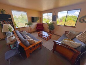 A living room with a sofa, armchair, coffee table, and a TV. There are plants, a lamp, and a bookshelf. - Ashland Commons Hostel (Ashland)