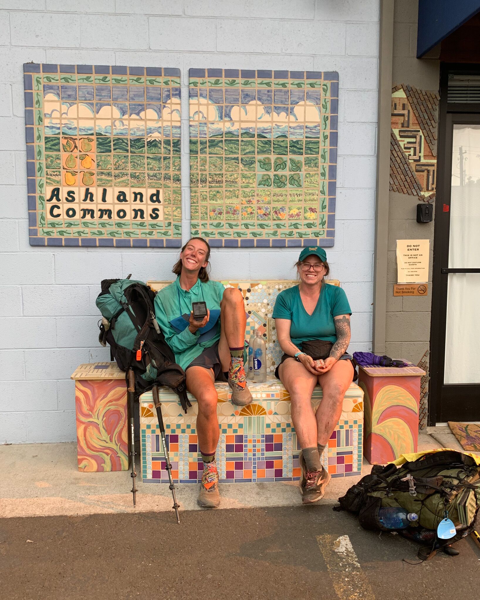 Two people sitting on a bench with backpacks, in front of a wall with a mosaic titled "Ashland Commons".