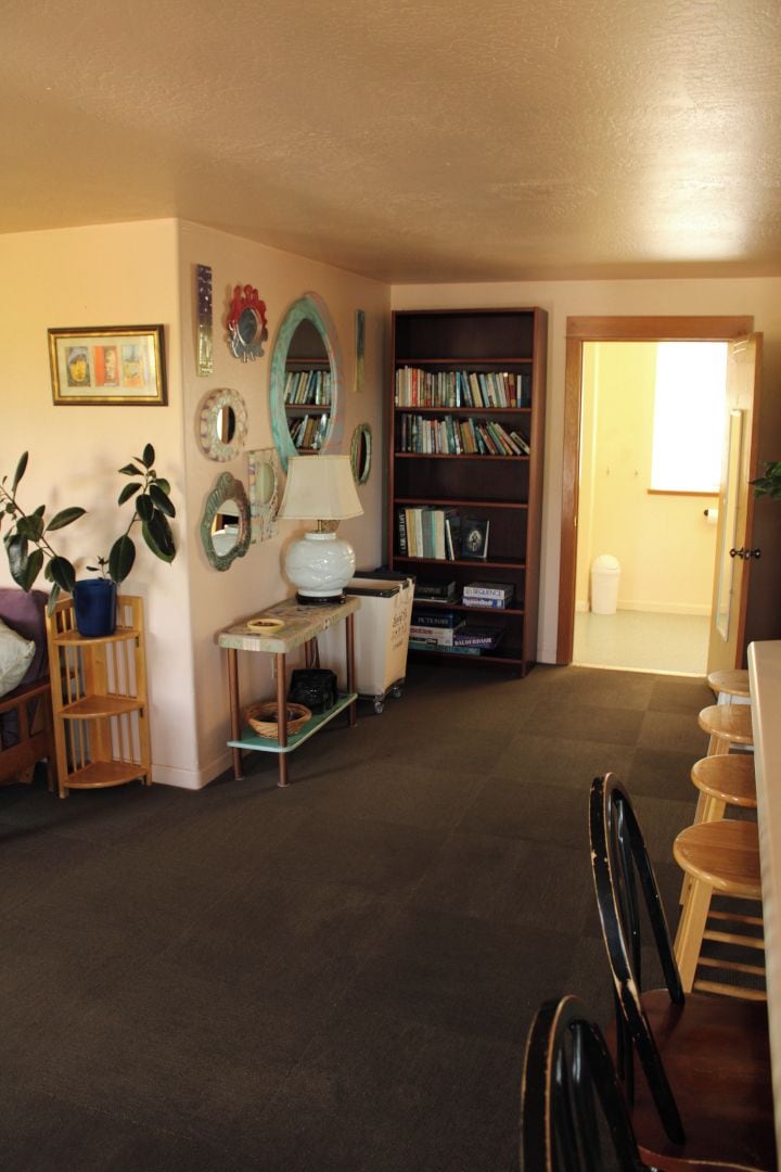 A living room with a bookshelf, a table with a lamp, and a door leading to another room.