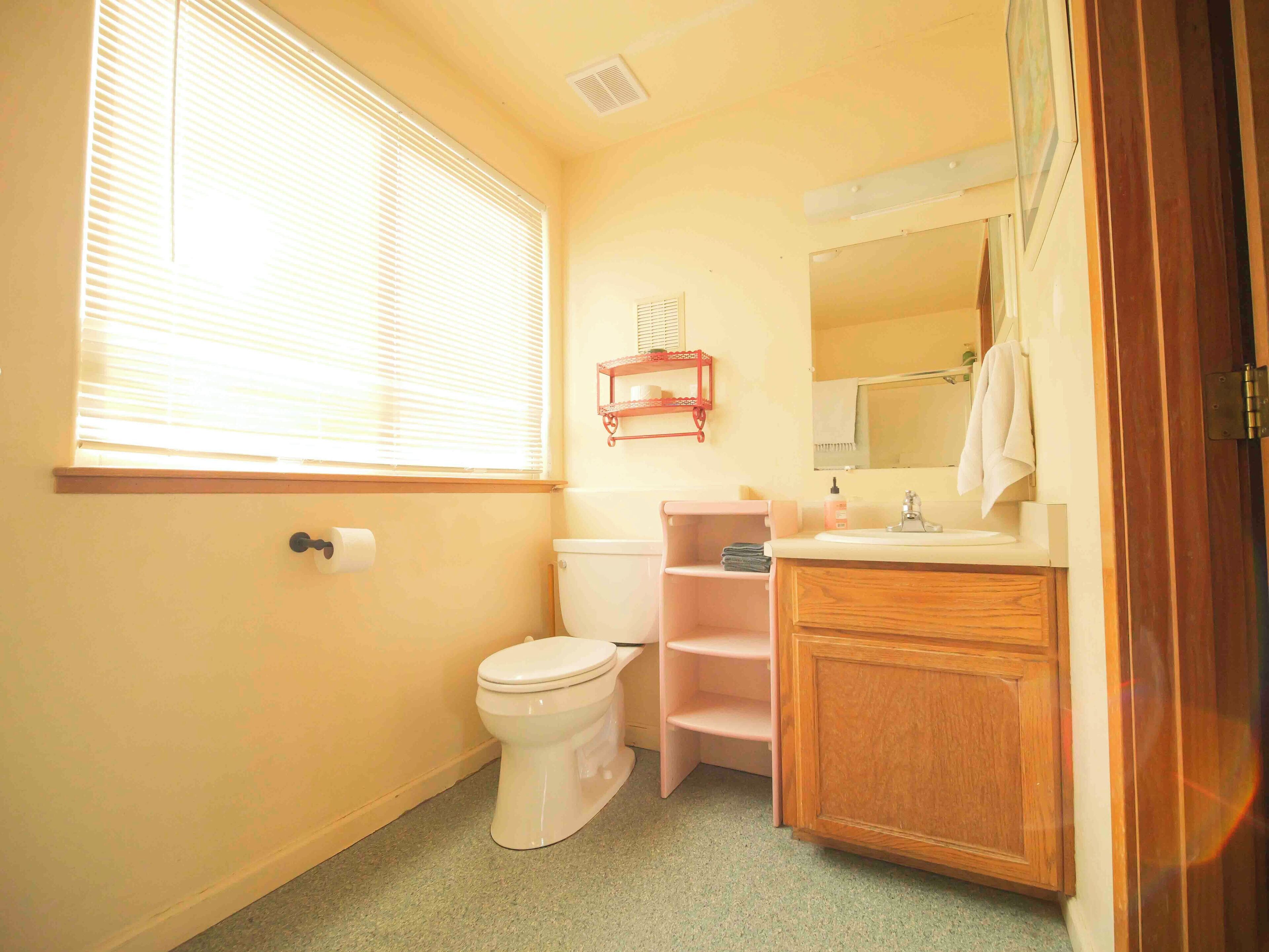 A bathroom with a toilet, sink, and vanity. There is a window with blinds, a mirror above the sink, and a shelf with toiletries.