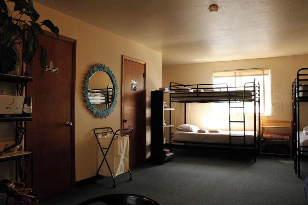 A dormitory room with bunk beds, a laundry cart, a rest room sign, and a window providing natural light.