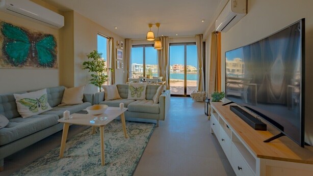 Ein modernes Wohnzimmer mit einer Couch, einem Couchtisch und einem Fernseher. Große Fenster bieten Blick auf das Meer.