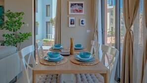 Ein Essbereich mit einem Holztisch, weißen Stühlen und blauen Tellern. An der Wand hängen gerahmte Bilder und es steht ein Topfpflanze.