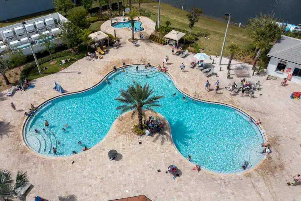 An aerial view of a swimming pool complex with multiple pools, surrounded by lounge areas and a grassy landscape.