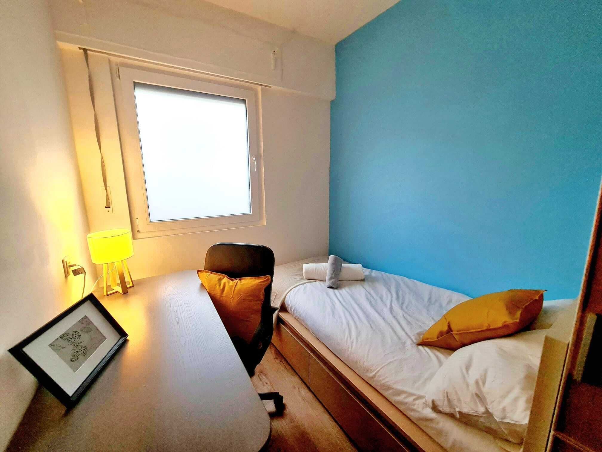 A small, well-lit room with a bed, a desk, a chair, and a window.